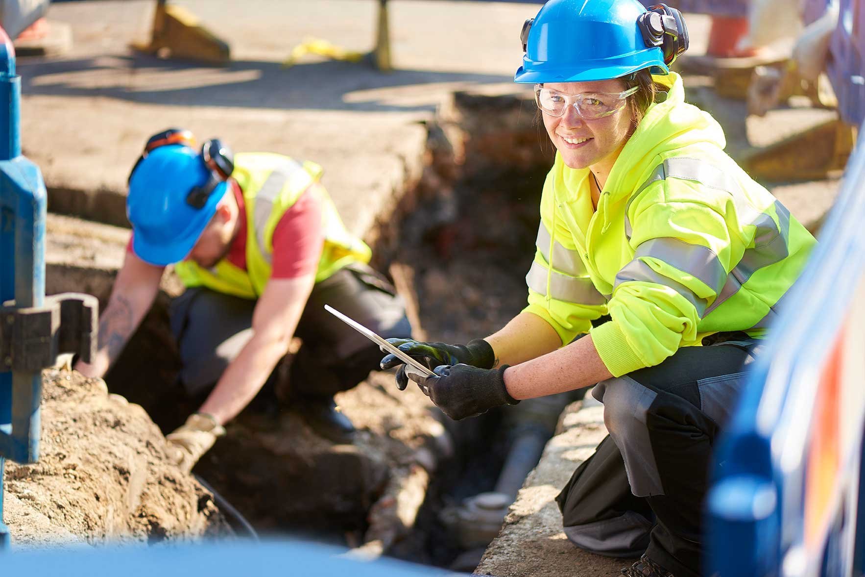 Junge Ingenieurin hockt am Rande einer Straßenbaustelle, im Hintergrund ist ein Arbeiter zu sehen.