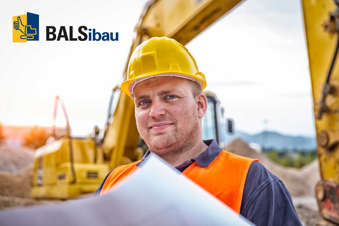 Ein Bauarbeiter mit gelbem Helm auf dem Kopf hält einen Plan in der Hand, hinter ihm ein Bagger